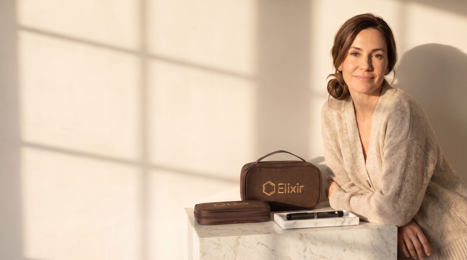 A woman resting against a marble plinth in window light, with the Elixir case, a smaller zip pouch and the pen set out in front of her